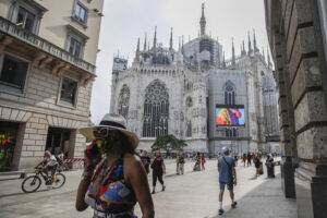 Pedestrians pass by a screen on the Duomo cathedral, showing a Moschino model during the Milan Digital Fashion Week, in Milan, Italy, Tuesday, July 14, 2020. Forty fashion houses are presenting previews of menswear looks for next spring and summer and pre-collections for women in digital formats, due to concerns generated by the COVID-19. (AP Photo/Luca Bruno)